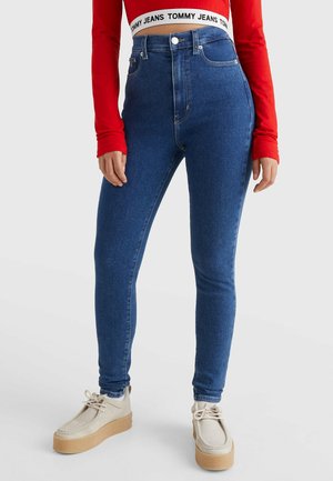 Femme portant un jean skinny bleu taille haute, un crop top rouge à manches longues, une ceinture blanche avec le texte "Tommy Jeans" et des chaussures compensées beiges.