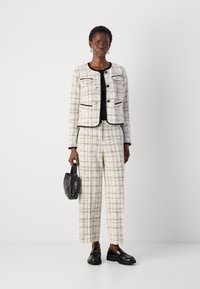 Ensemble de veste et pantalon en tweed crème avec un motif en grille et des accents noirs. Le modèle tient un petit sac à main tissé noir et porte des mocassins.