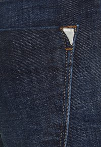 Pantalones vaqueros de mezclilla azul oscuro con una etiqueta triangular de marca de metal plateado en el bolsillo, costuras reforzadas y una superficie texturizada.