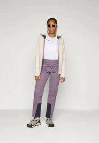 Naine, kellel on seljas prillid, valge lukuga jakk, valge T-särk, lilla püksid mustade paneelidega ja hallid matkajalatsid, seisab ühtlase tausta ees.