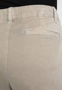 Pantaloni beige con una texture liscia, vestibilità slim e una tasca posteriore con apertura orizzontale. I punti di cucitura sono discreti e si abbinano al colore del tessuto.