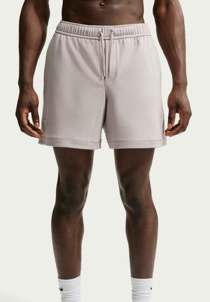Mann trägt hellgraue Sportshorts mit Kordelzug und weiße Sportsocken, steht mit locker an den Seiten hängenden Armen vor neutralem Hintergrund.