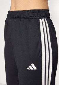 Svarta träningsbyxor i strukturerad tyg med vita sidosträngar och en Adidas-logotyp på nedre vänstra benet. Elastisk midja.