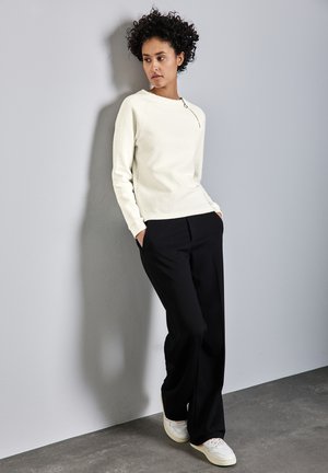 Weißer strukturierten Pullover mit einem Reißverschlussdetail, kombiniert mit schwarzen Hosen und weißen Sneakers. Das Modell posiert vor einer hellgrauen Wand.