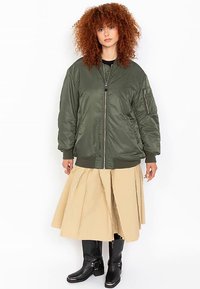 Veste bomber vert olive avec fermeture éclair à l'avant, poignets côtelés et poche latérale, portée sur une jupe évasée beige et des bottines noires.