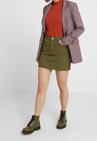 Blazer a cuadros en morado y blanco, suéter de cuello alto rojo brillante, falda de mezclilla verde oliva y botas de tobillo olive con cordones.