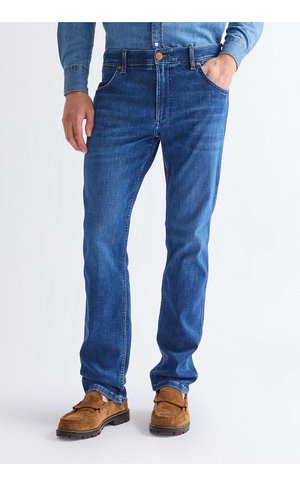 Muž nosící modré džíny, světle modrou džínovou košili a hnědé semišové mokasíny, s jednou rukou v kapse na bílém pozadí.