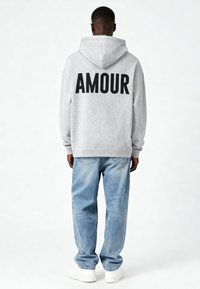 Šedá mikina s kapucňou s nápisom "AMOUR" vytlačeným tučnými čiernymi písmenami na zadnej strane, v kombinácii so svetlomodrými džínsami a bielymi teniskami.