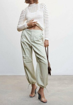 Kobieta w białym, dzianinowym, krótkim swetrze, beżowych spodniach ze sznurkiem, przezroczystych sandałach na wysokim obcasie, trzymająca brązową torebkę na pasku.