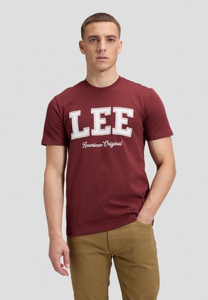 Junger Mann trägt ein dunkelrotes T-Shirt mit dem Text "LEE American Original" und Khakihosen, steht mit einer Hand in der Tasche vor schlichtem Hintergrund.