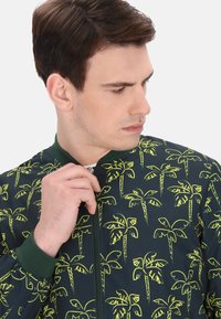 Groene bomberjack met een marineblauwe basis en gele palmboomprints, geribbelde kraag en manchetten, en een ritssluiting aan de voorkant.
