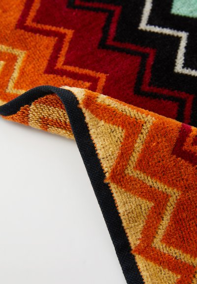 Tappeto colorato con un motivo a zigzag in rosso, arancione e nero. I bordi presentano una finitura scura con una superficie morbida e strutturata.