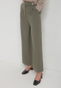 Pantaloni verde oliva a gamba larga con pince, caratterizzati da una chiusura a bottone. Abbinati a tacchi in vernice nera e a un maglione beige.