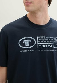 Tamsiai mėlynas medvilninis marškinėliai su ryškiu baltu spausdintu logotipu ir tekstu, kuriame nurodytos geografinės koordinatės, parašytos nusidėvėjusiame šriftu.