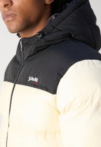 Giubbotto imbottito con parte superiore nera e parte inferiore crema, dotato di chiusura a zip, cappuccio e logo Schott sul petto. Tessuto liscio e isolante.
