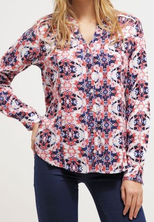 Femme portant un chemisier à manches longues boutonné avec un motif floral symétrique rouge, blanc et bleu marine, associé à un jean bleu foncé.