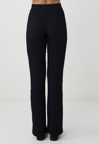 Une personne portant un pantalon noir à taille haute, droit et texturé, ainsi que des chaussures noires, se tenant devant un fond clair.