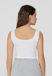 Weißes geripptes Crop-Top mit weitem Rundhalsausschnitt und breiten Trägern, kombiniert mit grauen Jogginghosen mit Kordelzug. Rückansicht zeigt eine glatte Textur und Passform.
