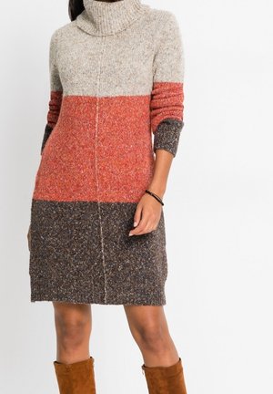 Donna che indossa un vestito a maglia a maniche lunghe a blocchi di colore beige, arancione e marrone, abbinato a stivali in camoscio marroni e un braccialetto nero.