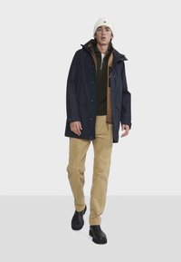 Marine waterdichte jas over een bruine ziptrui, gecombineerd met beige corduroy broek en zwarte laarzen; voorzien van een capuchon en meerdere zakken.