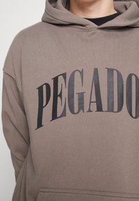 Felpa marrone con una grande grafica nera "PEGADO" stampata sul petto. Presenta un cappuccio con coulisse e una tasca a marsupio sul davanti.