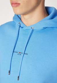 Felpa con cappuccio azzurro chiaro realizzata in misto cotone, con un logo frontale in nero, rosso e bianco. Include occhielli in tono argento e lacci coordinati.