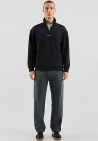 Schwarzer Hoodie mit Reißverschluss, hohem Kragen und weißem Logo auf der Brust, kombiniert mit lockeren dunkelgrauen Jeans und schwarzen Schuhen.