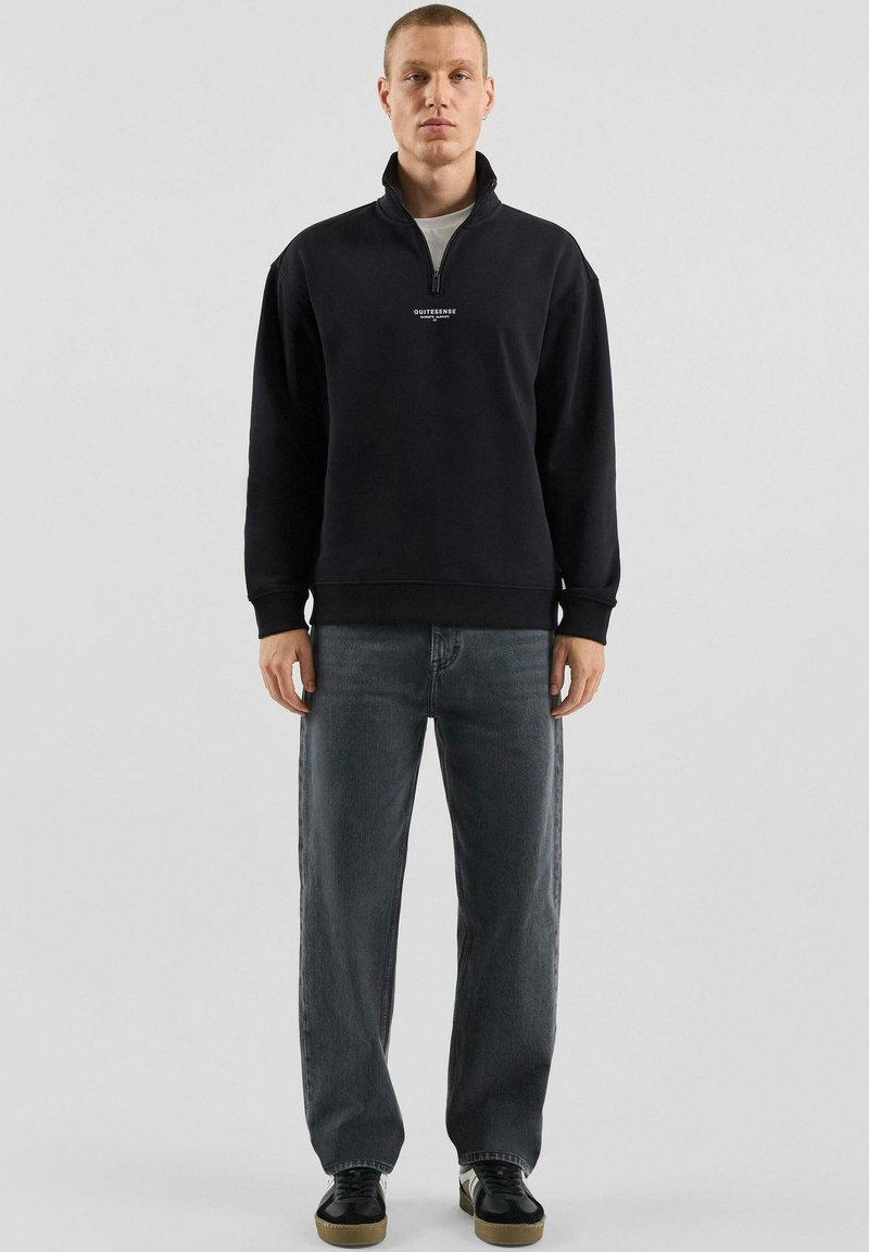 Schwarzer Hoodie mit Reißverschluss, hohem Kragen und weißem Logo auf der Brust, kombiniert mit lockeren dunkelgrauen Jeans und schwarzen Schuhen.