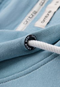 Jasnoniebieska dzianina z białym sznurkiem przewleczonym przez czarną metalową przelotkę z napisem „ombre wear”, na tle widoczne metki ubrania.