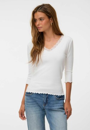 Femme aux longs cheveux bruns portant un haut blanc à col en V avec bordure en dentelle et un jean bleu, regardant légèrement sur le côté.