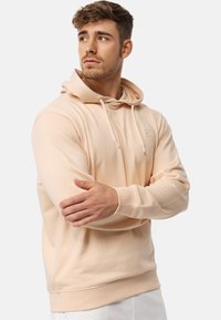 Sudadera de color melocotón hecha de un tejido suave, con capucha ajustable, puños acanalados y un pequeño logo en el pecho. Textura lisa.