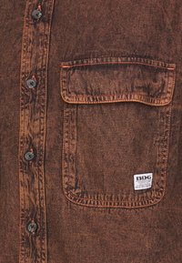 Brun denimskjorta med en framficka, svarta knappar och kontrasterande orange sömmar. Tagg synlig på fickan med varumärkesnamn.