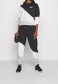 Svart och grå idrottshoodie med en kortad design, vita detaljer och matchande sweatsbyxor med ett böjt mönster; modellen håller i en viktskiva.