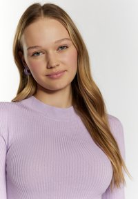 Lila bordázott, hosszú ujjú blúz magas gallérral. A modellen kerek, lila fülbevalók láthatók. Hosszú, hullámos haja semleges arckifejezést keretez.