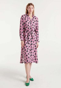 Vestido rosa con mangas largas y un cinturón, con un patrón de fresas negras y verdes. Lleva zapatos planos verdes.