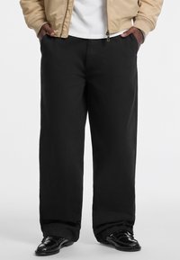 Black wide-leg pants made from a smooth fabric, paired with a beige bomber jacket and black shoes, showcasing a relaxed, casual fit.