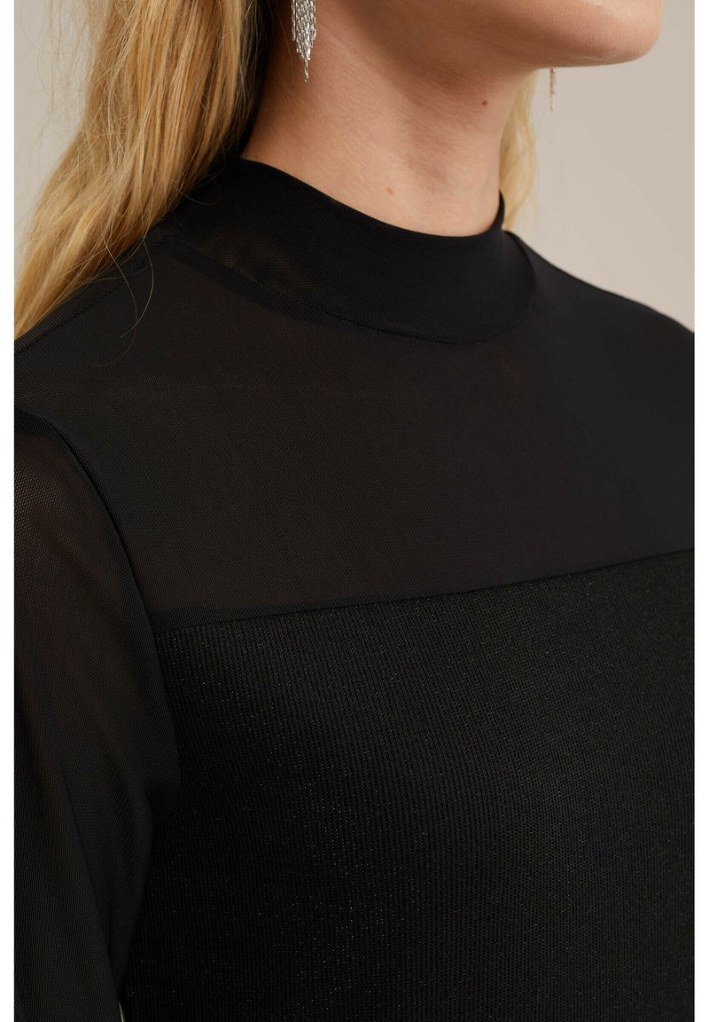 Zwarte top met een hoge halslijn, voorzien van een doorschijnend bovenstuk en een gestructureerde stof eronder. Geaccentueerd met een glinsterend detail bij de hals.