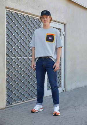Camisa cinza de mangas curtas com um bolso colorido, jeans escuros e ténis brancos com detalhes em laranja. Fundo urbano com rede metálica.