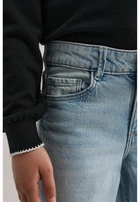 Käsi toetub helerohelise teksapükste tasku kõrvale, kannab musta kampsunit valge mansetiäärisega.