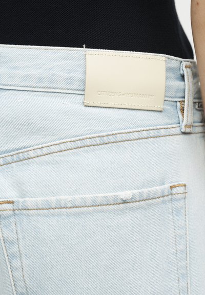 Ανοιχτό μπλε τζιν με ετικέτα από μπεζ δέρμα ραμμένη πάνω από την πίσω τσέπη, φορεμένα με μαύρο τοπ.