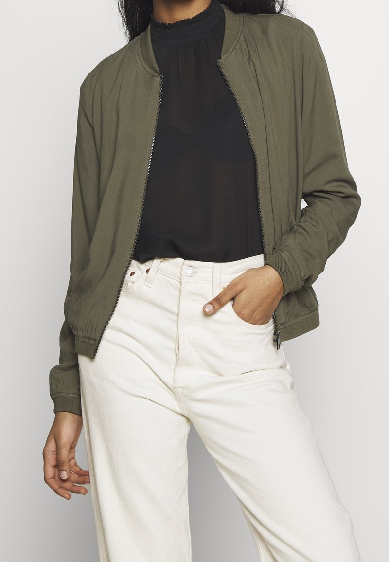 Olive green bomber jacket with ribbed cuffs and collar; paired with a sheer black top and white high-waisted pants; zipper detail visible.