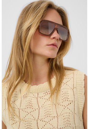 Hosszú, szőke hajú nő, nagy barna négyzet alakú napszemüveget visel és mintás krémszínű kötött ujjatlan felsőt.