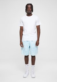 Hvid t-shirt, lyseblå tie-dye shorts, hvide sneakers og hvide sokker med sort logo. Modellen står foran en ensfarvet baggrund.
