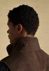 Hnedá fleecová vesta s vysokým golierom, s vyrazeným logom "JOULE" na chrbte, hladká textúra a kontrastné švy.