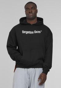 Black hooded sweatshirt with "forgotten faces" white text, front pocket, and ribbed cuffs. Model wears light gray pants.