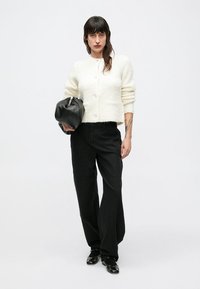 Witte, gestructureerde, cropped cardigan met knopen; gecombineerd met hoog getailleerde zwarte jeans en een zwarte tas. Schoeisel is zwart, glanzend, platte schoenen.