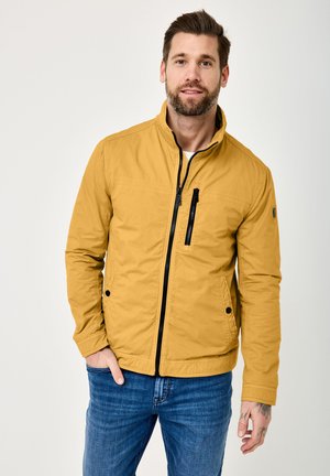 Man with beard wearing mustard yellow zip-up jacket and blue jeans, standing with one hand in pocket against plain background.
