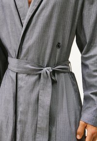 Bata gris en una tela texturizada con cinturón de lazo, con ribete en negro contrastante y un logo en el área del pecho izquierdo.