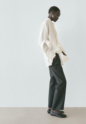 Frau in weißem, übergroßem Blazer und schwarzen Hosen mit weitem Bein, steht seitlich mit den Händen in den Taschen vor einem schlichten Hintergrund.