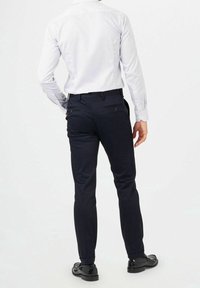 Heren nauwsluitende marineblauwe broek met een strakke uitstraling, twee achterzakken en een slim fit. G gedragen met een licht gestreept overhemd met lange mouwen en schoenen.